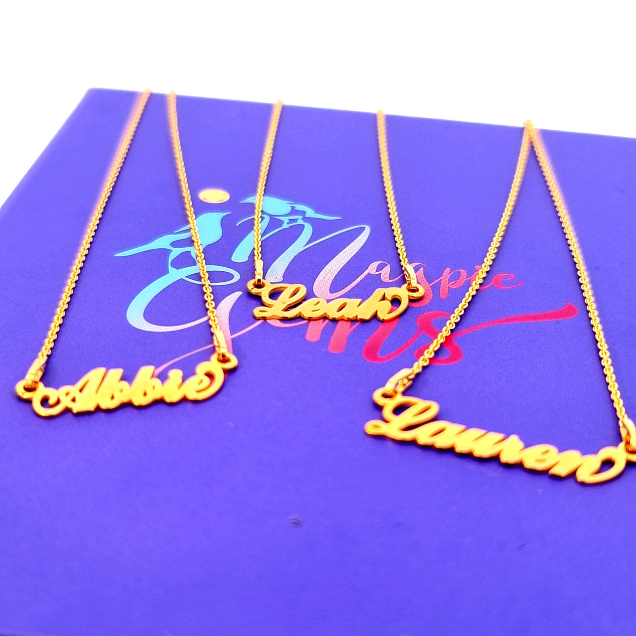 Close-up of personalised gold name necklaces with names "Abbie," "Leah," and "Lauren," displayed on a purple Magpie Gems branded background.