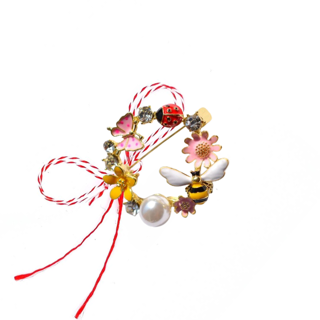 Spring Garden Martisor Brooch on a white background, featuring a circular design adorned with enamelled flowers, a bee, a ladybird, a butterfly, sparkling crystals, and a central pearl, paired with the traditional red and white Martisor bow to symbolise spring and vitality.